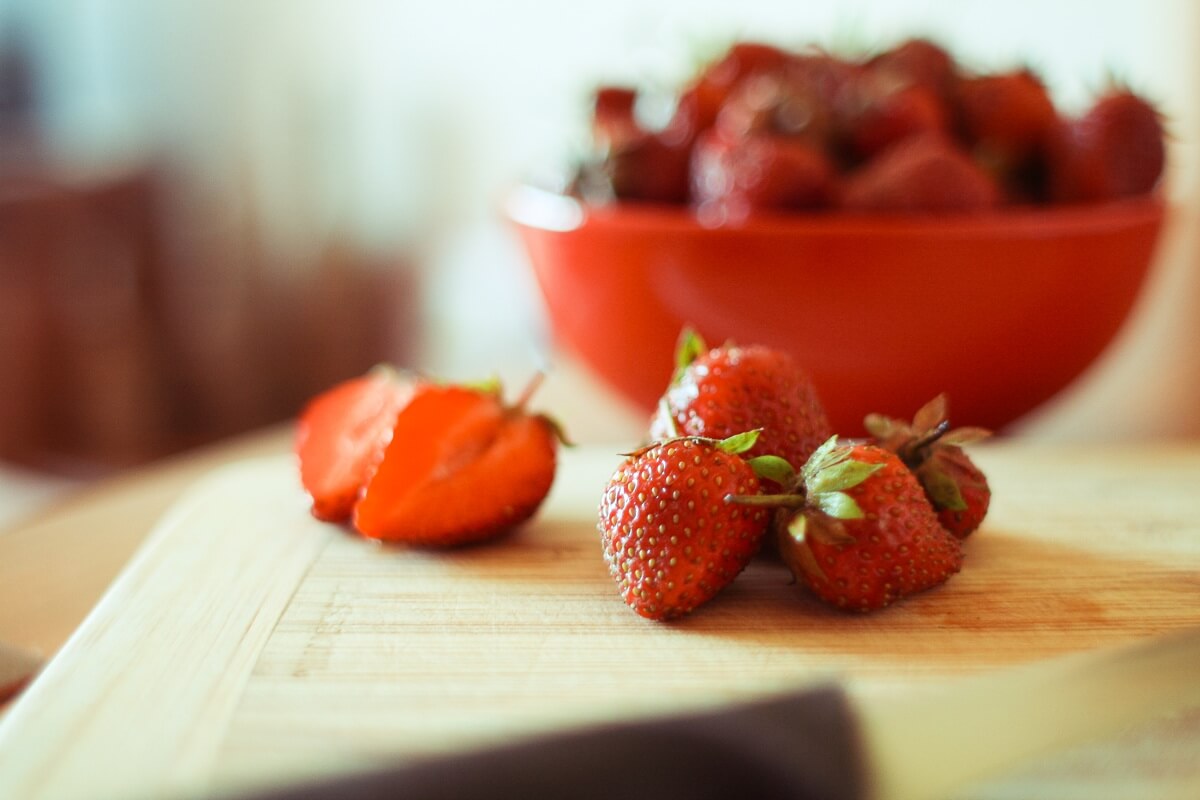 Auf einem hölzernen Küchenbrett liegen einige Erdbeeren vor einer roten Schale mit Erdbeeren