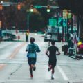 Two runners are seen from behind running down a major road in a city.