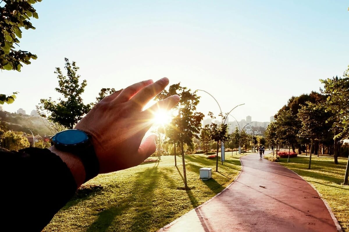 A hand wearing a smartwatch shields the low sun over a park path, with runners in the distance.