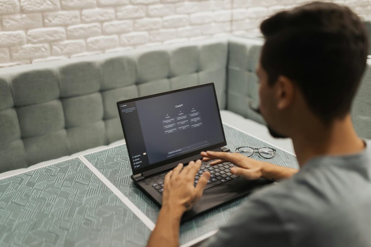 Person sitting at a table using a laptop with the ChatGPT interface open on the screen; a pair of glasses rests beside the laptop in a cushioned booth.