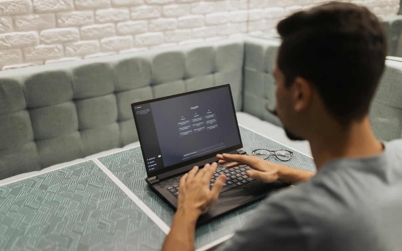 Person sitting at a table using a laptop with the ChatGPT interface open on the screen; a pair of glasses rests beside the laptop in a cushioned booth.