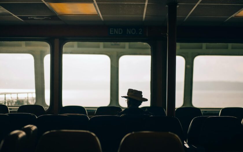 Silhouette of a passenger in a brimmed hat sitting among empty seats inside a ferry, with bright windows behind.