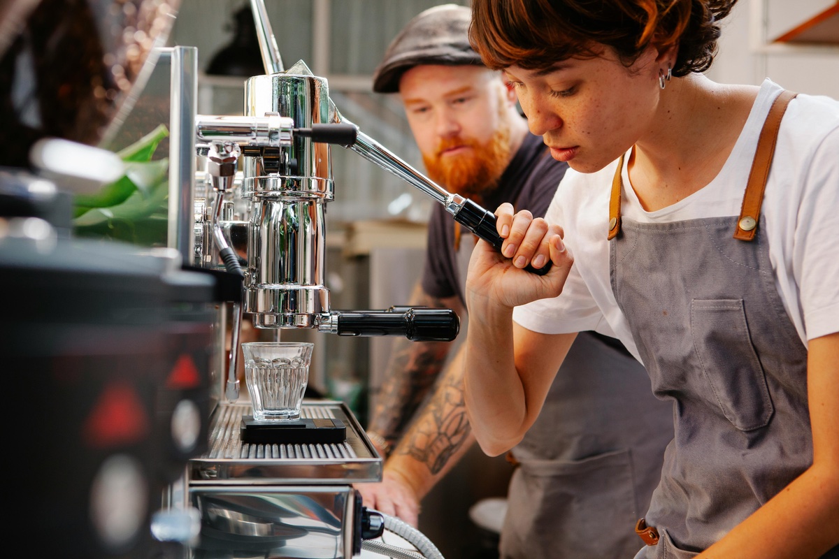 A barista carefully pulls a lever on a chrome espresso machine while preparing a shot of coffee, as a colleague watches in the background inside a café.