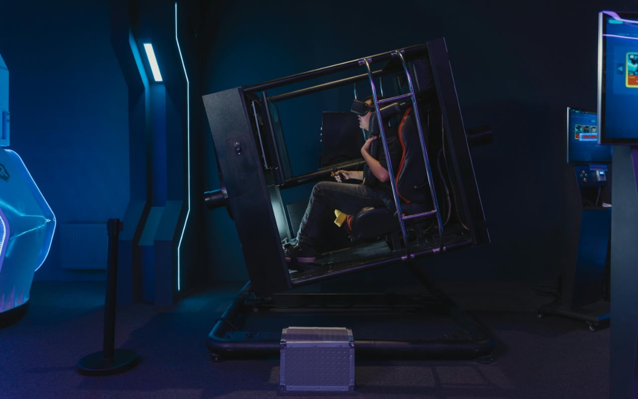 Person wearing a VR headset seated in a tilted motion-simulator cockpit inside a blue-lit arcade room.