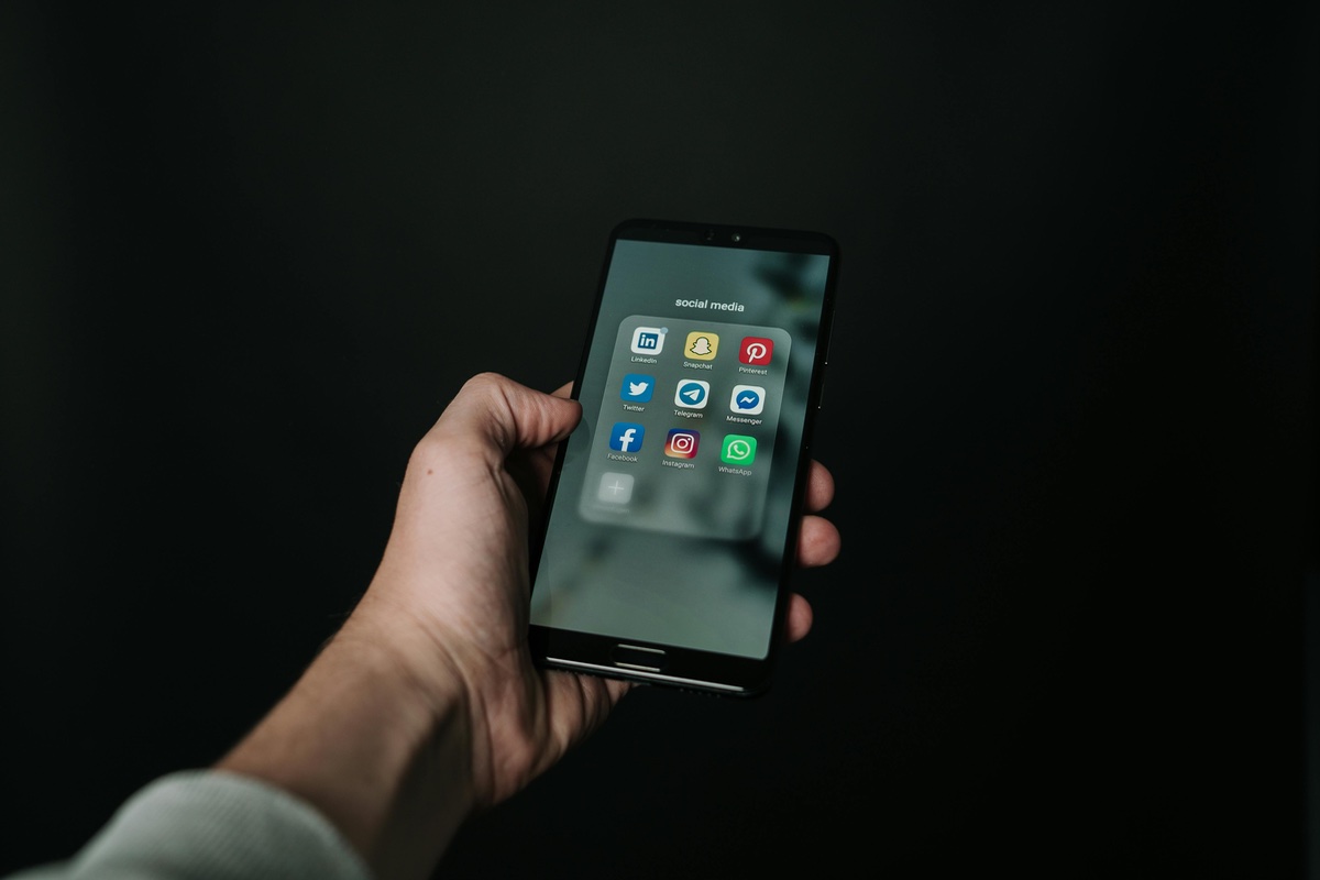 Hand holding a smartphone against a dark background, displaying a folder of social media apps including LinkedIn, Snapchat, Pinterest, Twitter, Telegram, Messenger, Facebook, Instagram, and WhatsApp.