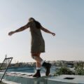 A tattooed person in a short dress balances with outstretched arms while walking along the edge of a rooftop, with city buildings and a clear sky in the background.