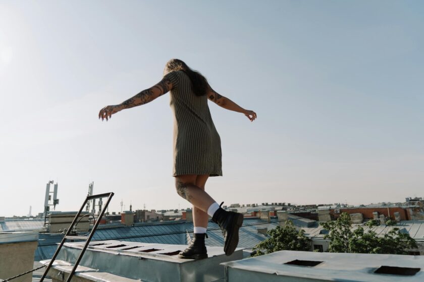 A tattooed person in a short dress balances with outstretched arms while walking along the edge of a rooftop, with city buildings and a clear sky in the background.
