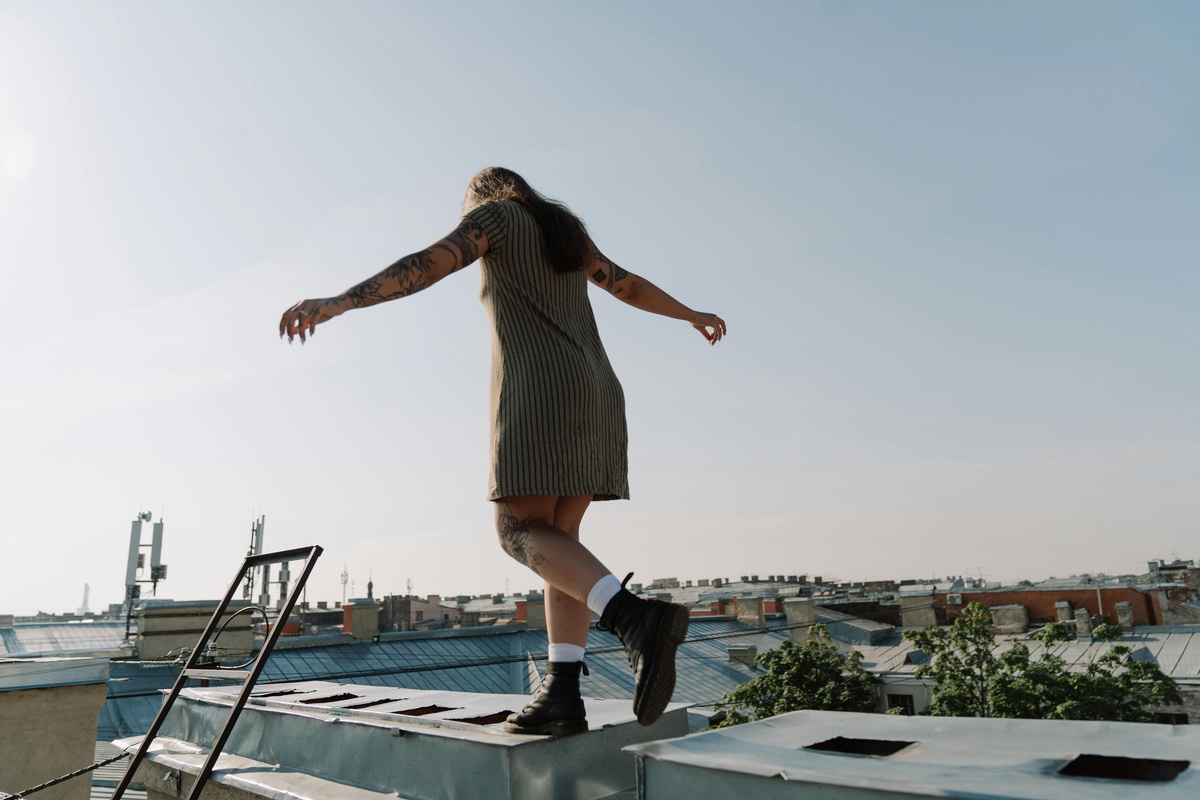 A tattooed person in a short dress balances with outstretched arms while walking along the edge of a rooftop, with city buildings and a clear sky in the background.