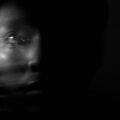 A blurred black-and-white image of a person’s face emerging from darkness, with motion creating a ghost-like double exposure effect.
