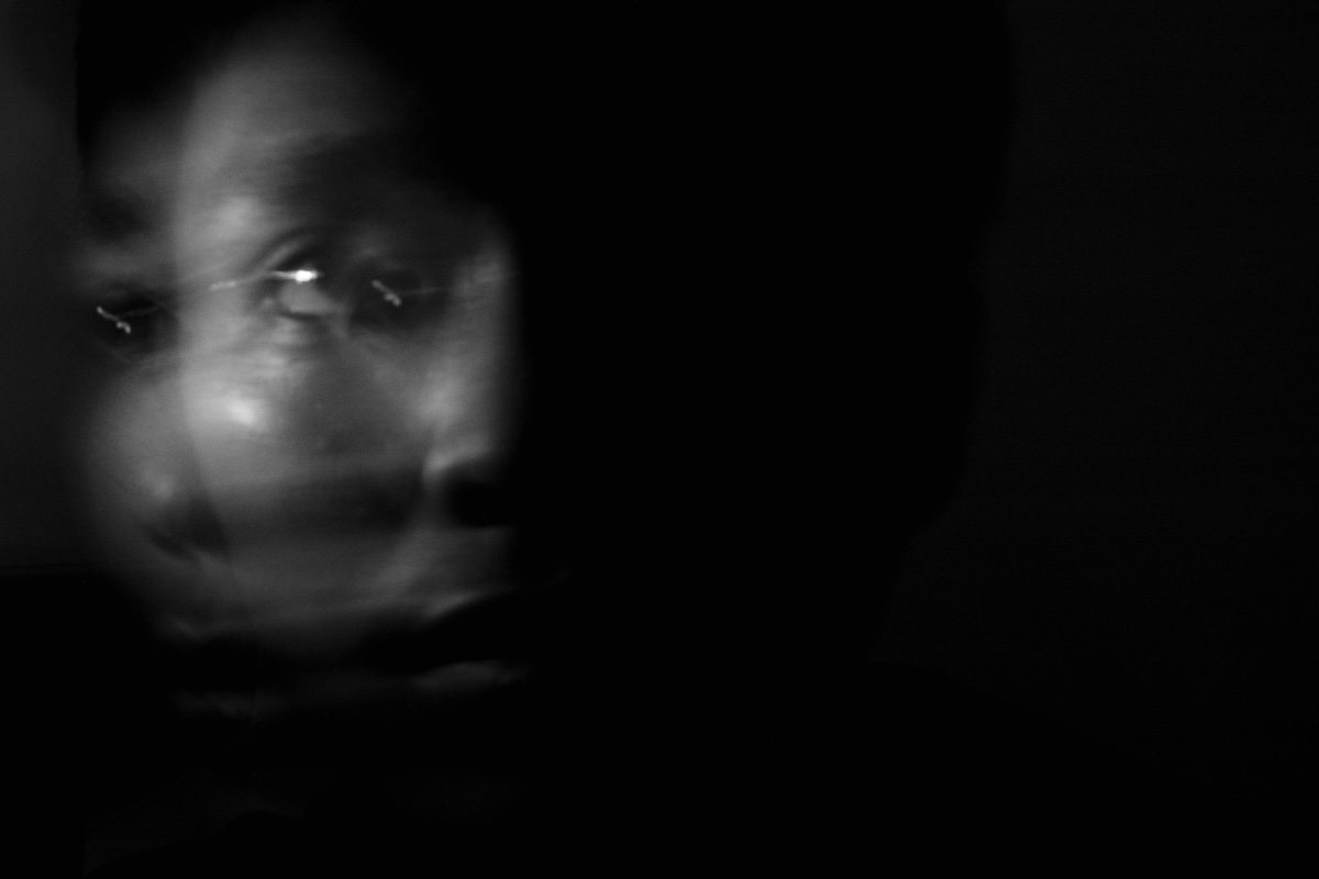 A blurred black-and-white image of a person’s face emerging from darkness, with motion creating a ghost-like double exposure effect.