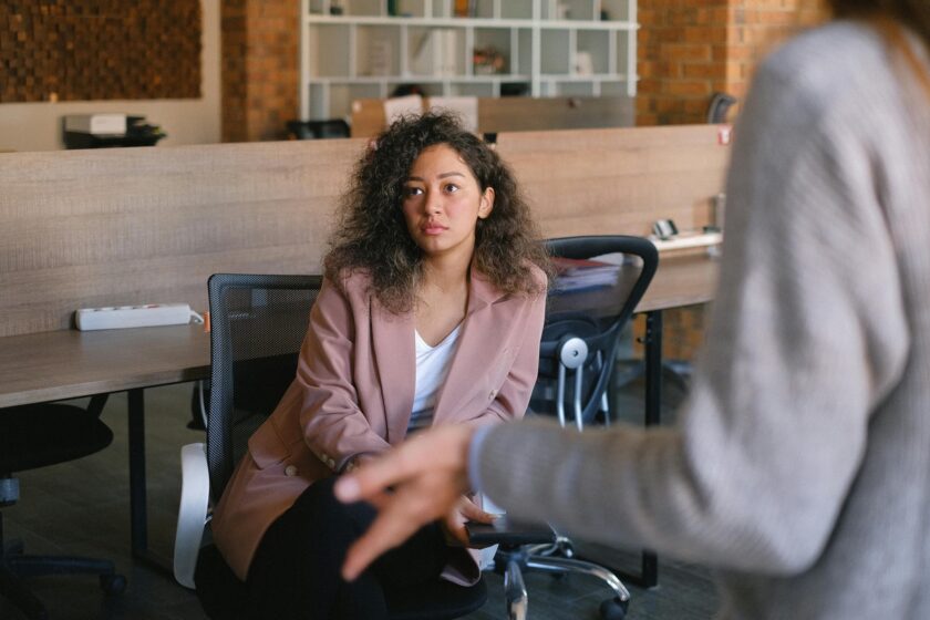 A woman sits in an office chair listening attentively during a conversation, while another person gestures in the foreground in a modern workspace.