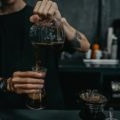 A person pours freshly brewed coffee from a glass carafe into a tall glass while preparing a pour-over at a café counter.