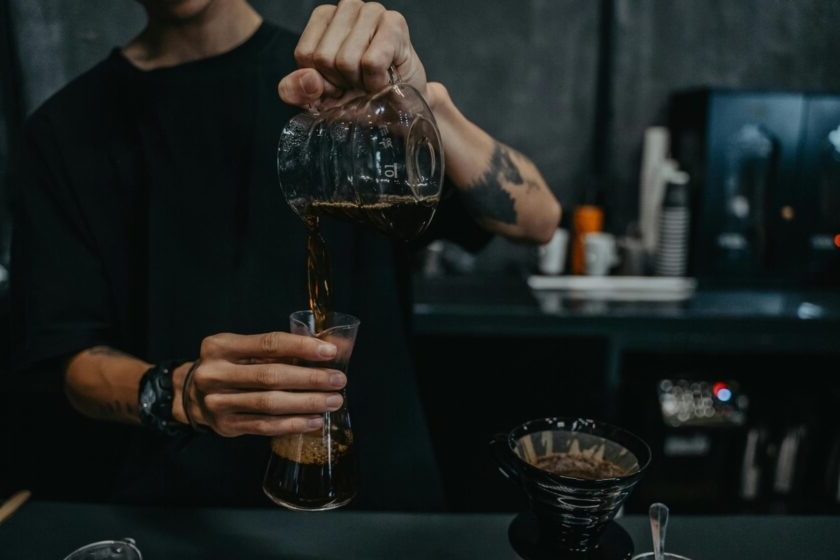 A person pours freshly brewed coffee from a glass carafe into a tall glass while preparing a pour-over at a café counter.