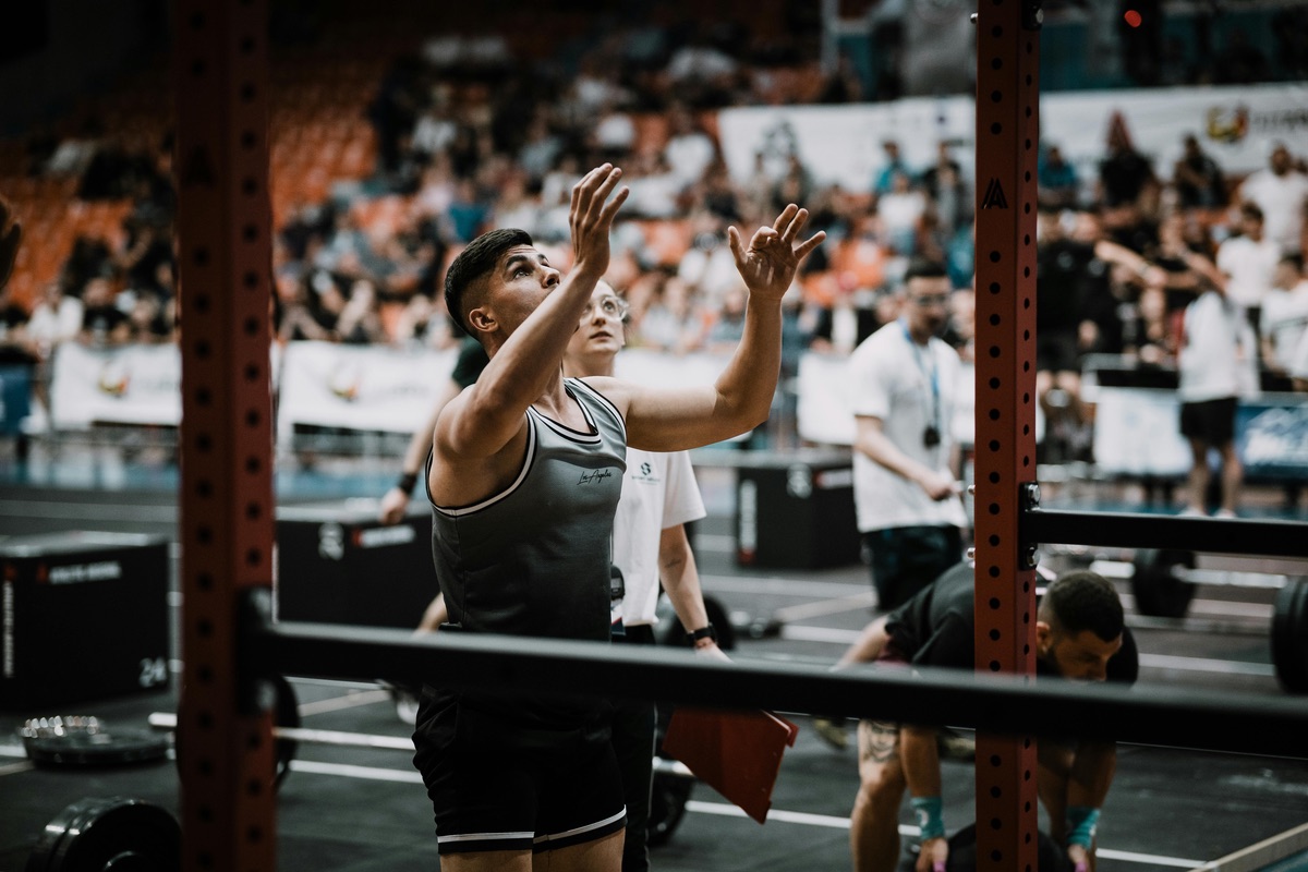 A male athlete reaches upward during a fitness competition inside a stadium, with judges and other competitors visible in the background.