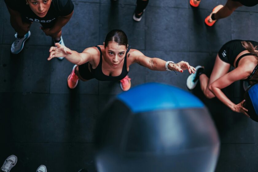 Overhead view of a woman in a gym reaching up to catch or push a large medicine ball, surrounded by other participants during a group workout.