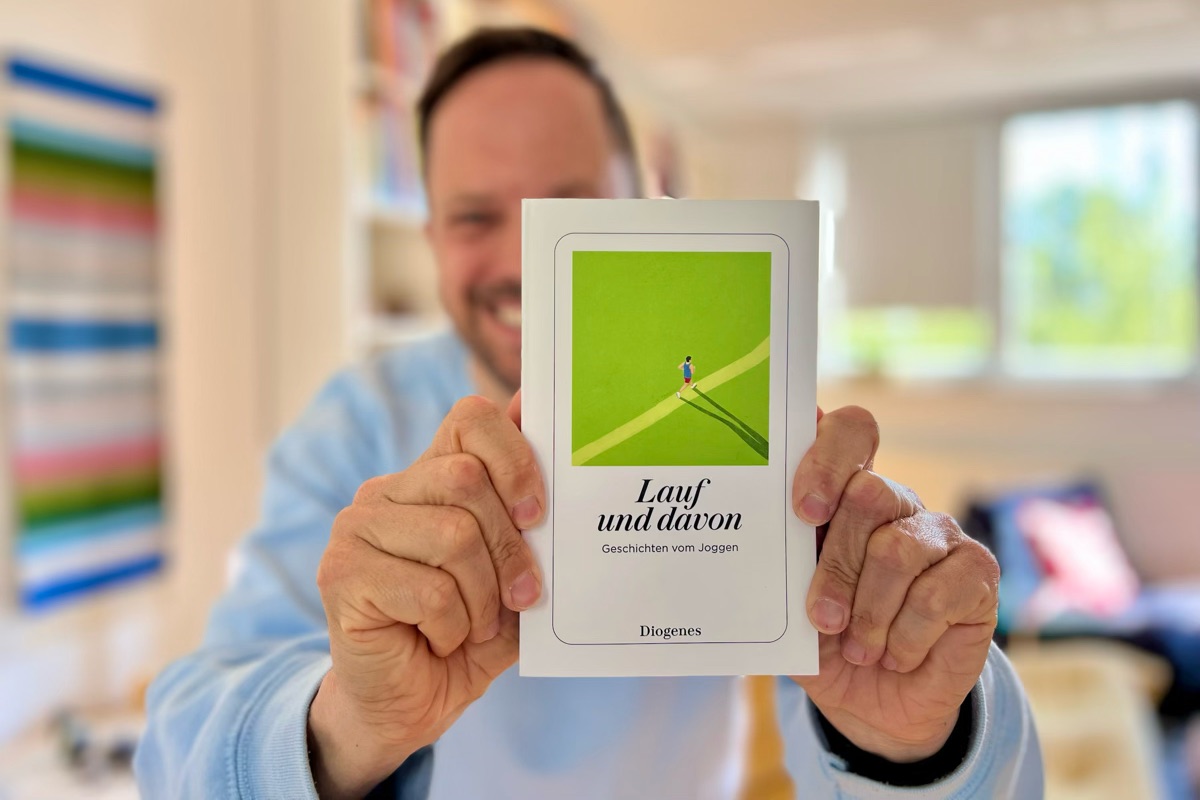 A smiling person holds up a book titled "Lauf und davon: Geschichten vom Joggen" by Diogenes, with the cover showing a runner on a green path.