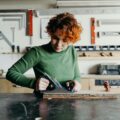 A person uses a hand plane to smooth a piece of wood on a workbench in a well-organized workshop with tools mounted on the wall.