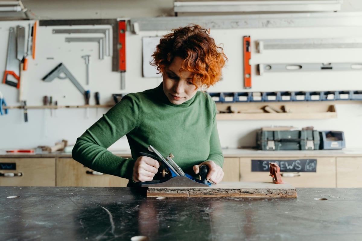 A person uses a hand plane to smooth a piece of wood on a workbench in a well-organized workshop with tools mounted on the wall.