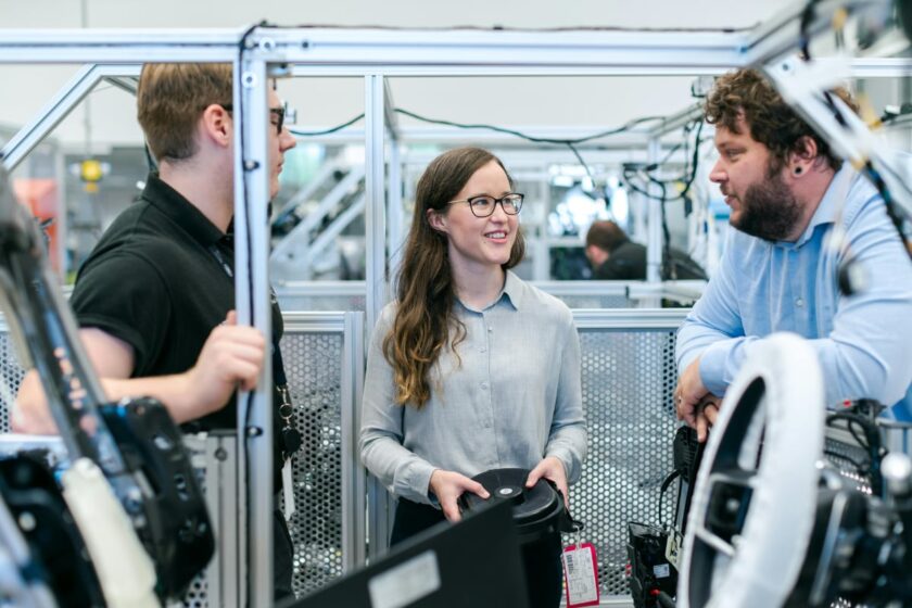 Three colleagues stand together in an industrial workspace, discussing equipment while one person holds a component and the others listen attentively.