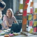 Two women collaborate at a desk with a laptop in a bright workspace, surrounded by colorful sticky notes on a glass wall.