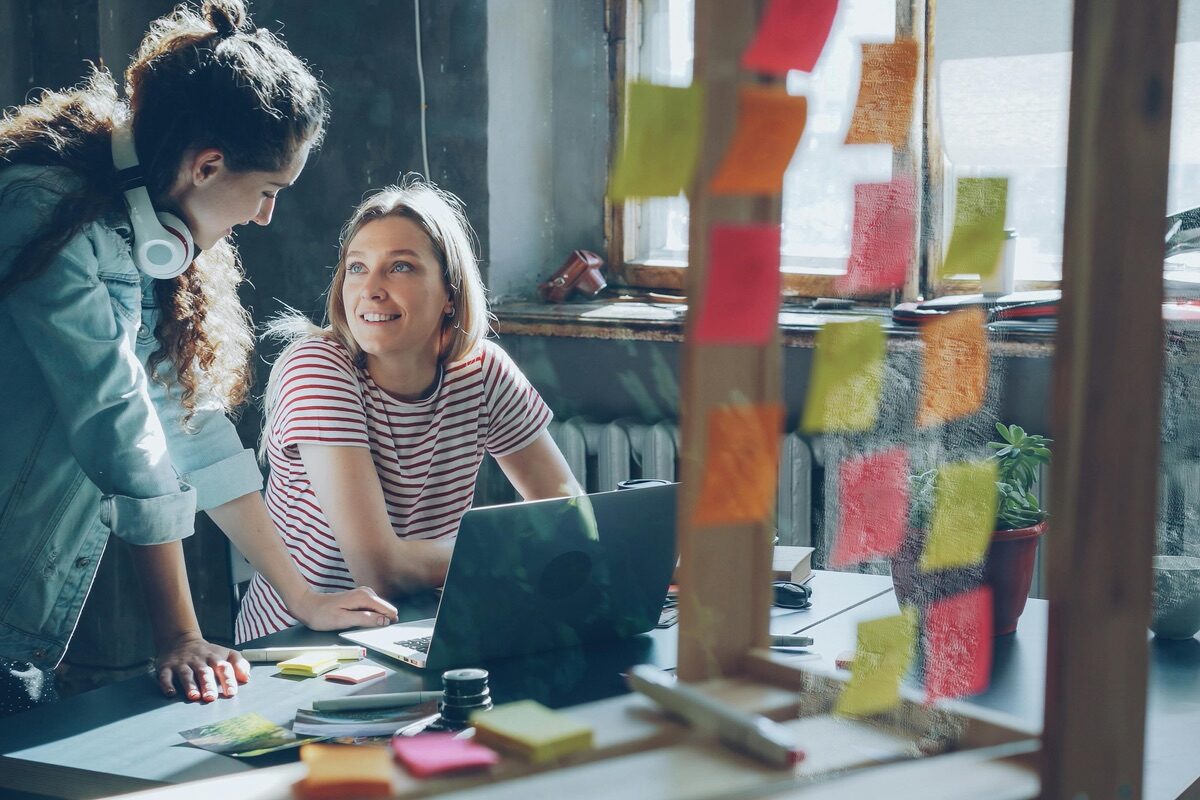 Two women collaborate at a desk with a laptop in a bright workspace, surrounded by colorful sticky notes on a glass wall.
