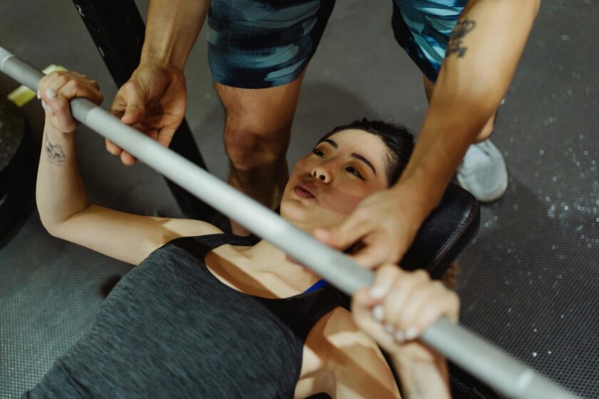 A person lies on a workout bench gripping a barbell while another person assists during a weight training session at the gym.