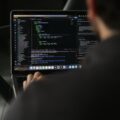 A person works on a laptop displaying source code and development tools, viewed over the shoulder in a dimly lit workspace.