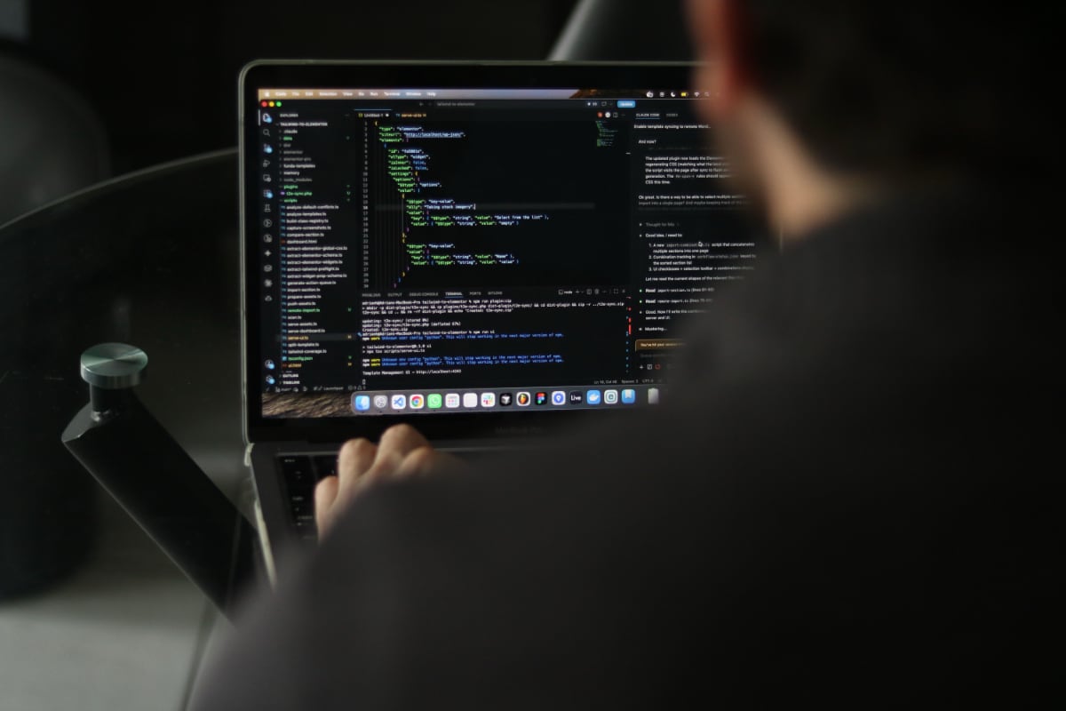 A person works on a laptop displaying source code and development tools, viewed over the shoulder in a dimly lit workspace.