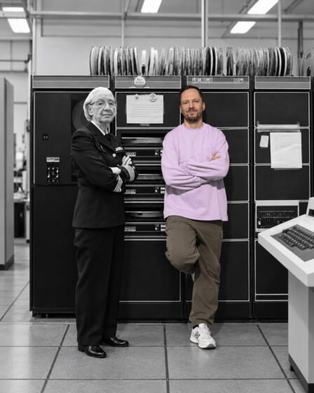 AI-generated image of two people standing with folded arms in front of a large vintage computer system with rows of magnetic tape reels. The scene is mostly black and white, while one person’s pink sweatshirt and beige trousers are shown in color.