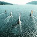 An aerial view of several sailboats racing across open water, leaving white wakes behind them, with mountains framing the horizon.