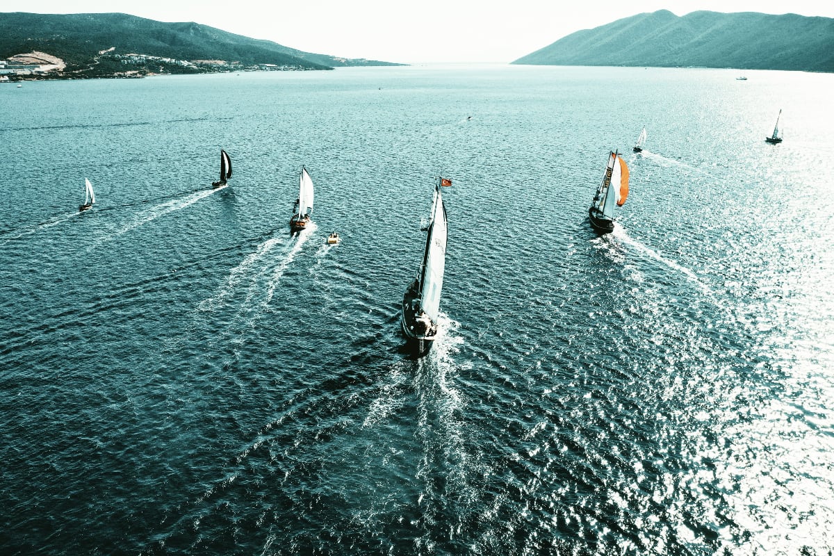 An aerial view of several sailboats racing across open water, leaving white wakes behind them, with mountains framing the horizon.